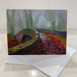 13.9 x10.7 card. With envelope. Image of autumnal Oldbridge canal bridge with bare trees in background and fallen copper red and purple shadowed leaves on ground in front,  image of an original painting.  Blank inside card