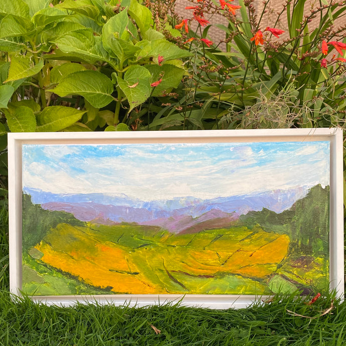 yellow fields in foreground of abstracted landscape painting   mountains fade into the background   image shows the painting resting against shrubbery in garden outdoors