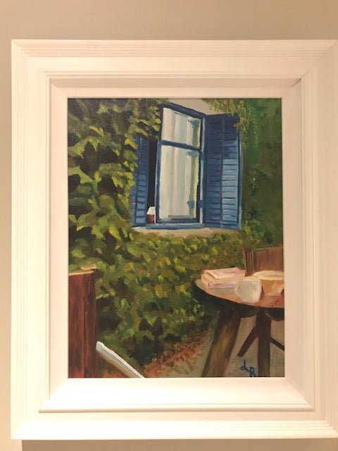 white framed painting showing an ivy covered wall with a blue shuttered window open, round table with books and coffee cup and two chairs   summer vibes   holiday home